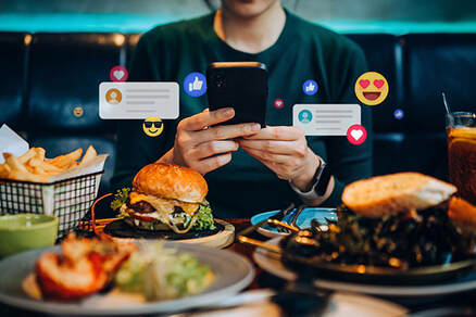 This image shows a patron at restaurant table with several dishes in the foreground, including a cheeseburger on a plate and a basket of fries. Cropped at the chin, the photo shows only the neck, torso, arms, and hands of the patron, who is wearing a black t-shirt and typing on a black smartphone. Superimposed on the photo are assorted emojis, suggesting the patron has posted on social media and is getting “likes” and other feedback. Insurance underwriters look at what your customers are posting for clues about the nature of your business. A mismatch between your insurance application and online reputation can result in an adverse underwriting decision. 